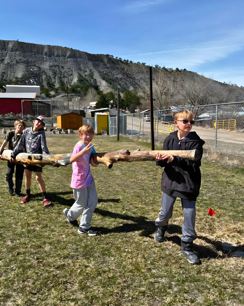 Students carrying a large log together outdoors on a grassy field, working as a team with mountains in the background.