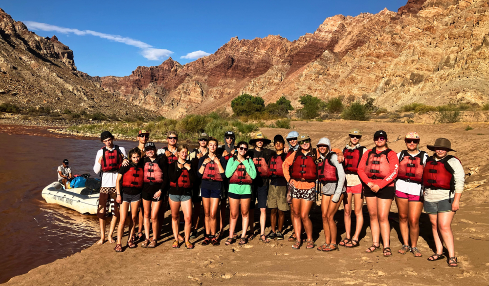 Class of 2026 students in red life jackets on Cataract Canyon shore with rafts and cliffs behind them