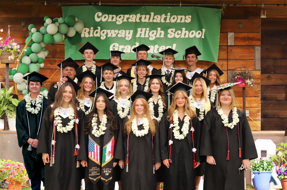 Group photo of Ridgway High School’s Class of 2026 graduates in caps and gowns standing in front of a green congratulatory banner at graduation.