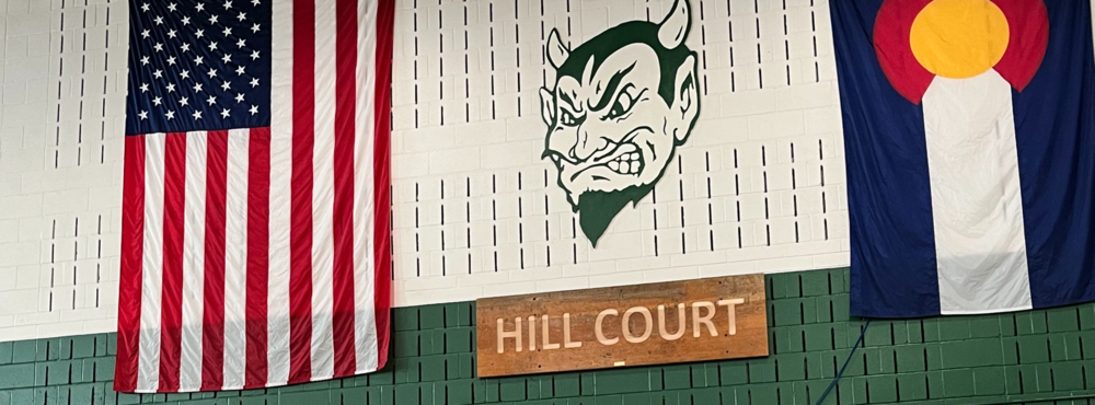 Gym wall with large American and Colorado flags hanging beside a green devil mascot logo above a wooden “Hill Court” sign.