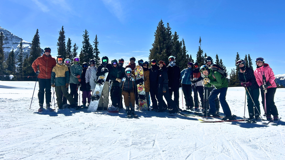 Group of high school students and adults in ski gear standing together on a snowy mountain with trees and blue sky in the background.