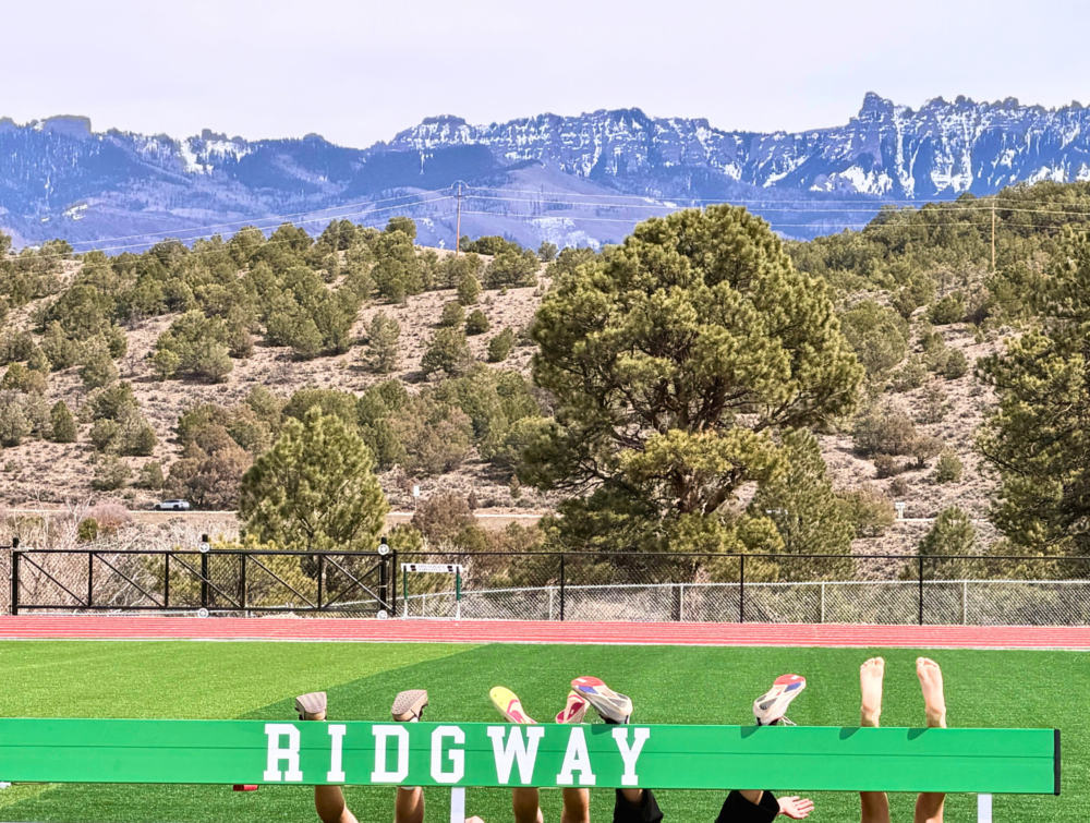 Athletes’ legs and shoes stick up from behind a green hurdle labeled “RIDGWAY,” with a track, trees, and snow-capped mountains in the background.