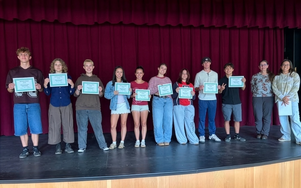 Nine high school students stand on a stage holding certificates, with two adults beside them, in front of red theater curtains.