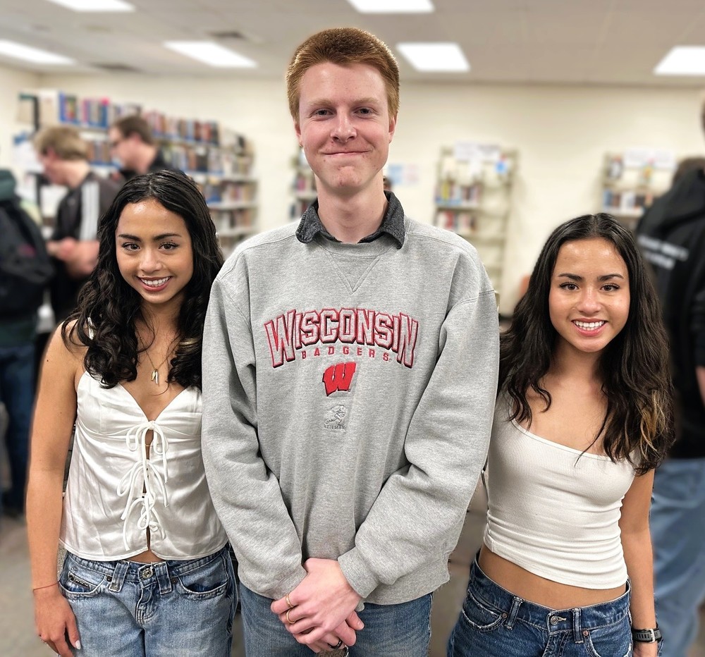 Three students standing indoors, smiling at the camera, with bookshelves and other people visible in the background