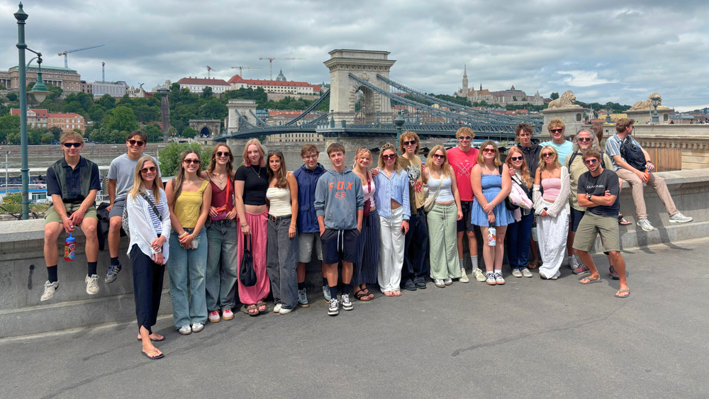 Students and graduates posing for a group photo in Budapest with the Chain Bridge and city skyline in the background under cloudy skies.