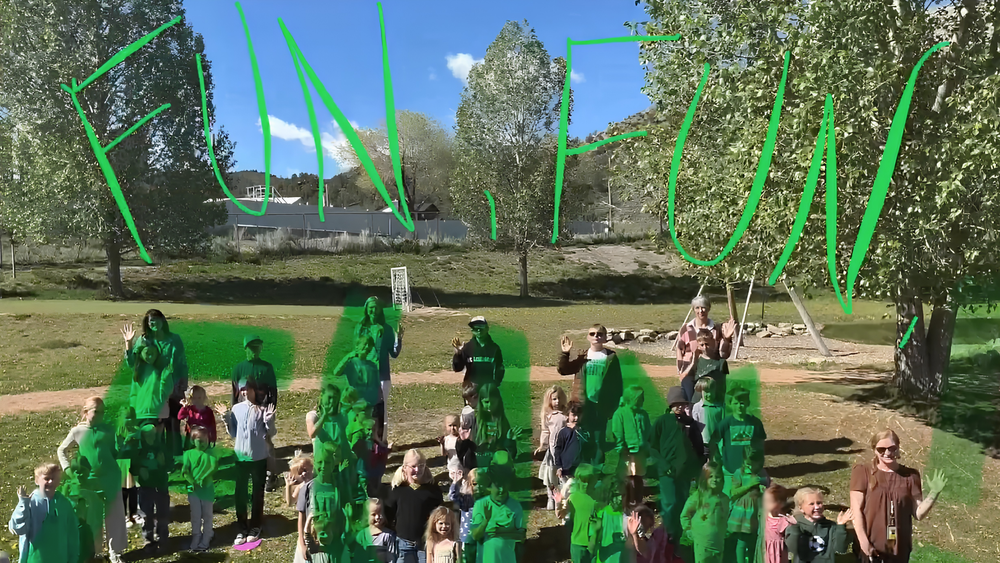 A group of children and adults are gathered outside on a grassy field, waving at the camera. The group is standing in front of a row of trees, with a clear blue sky above. The word "FUN" is written in large green letters across the top of the image, partially covering the people.ritten above..