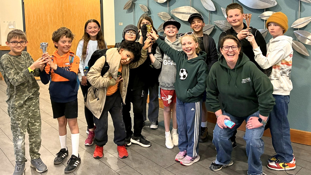 Group of students and one adult smiling and posing indoors, holding small trophies and celebrating a math competition achievement.