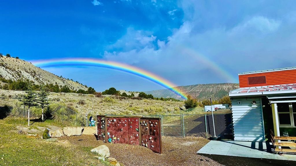 A vibrant double rainbow arcs across a bright blue sky above grassy hills and a school playground, with two children near a small climbing wall and building.