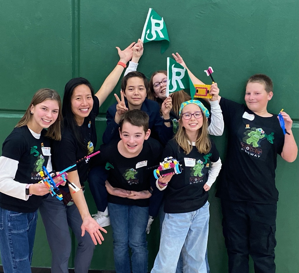 Group of students and coach in front of green wall holding flags and lego robots.