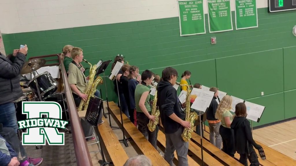 Pep band students standing on gym bleachers playing instruments during a game, with green walls and championship banners in the background.