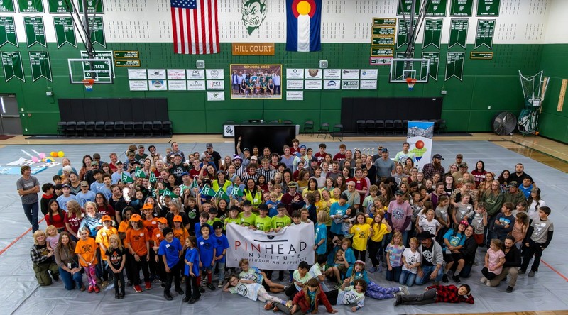 A large group of students, coaches, volunteers, and families gathered in a school gym for a group photo after a robotics tournament, with colorful team shirts and a Pinhead Institute banner at the front.