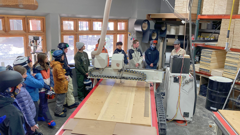 High school students standing inside a ski manufacturing workshop while listening to a staff member explain equipment and ski production.