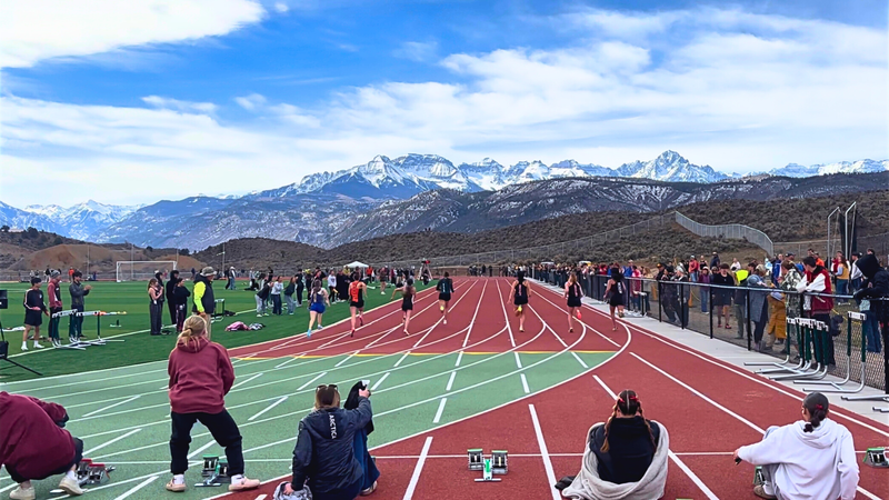 Runners sprint down a track during a meet, with spectators lining the sides and snow-covered mountains in the background.
