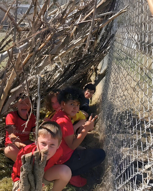Group of students sitting together inside a small stick shelter next to a fence, smiling and gathered closely during an outdoor activity.