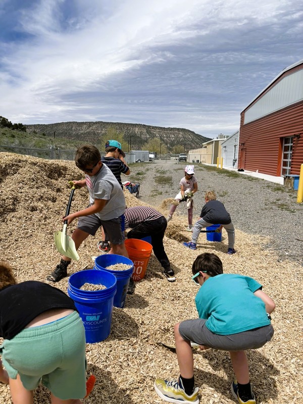 Students use buckets and shovels to move wood chips outside near a playground on a sunny day.
