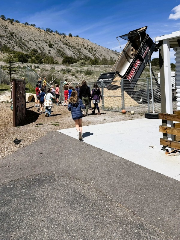 Students gather outside to watch a dump truck unload wood chips near a playground, with hills and blue sky in the background.
