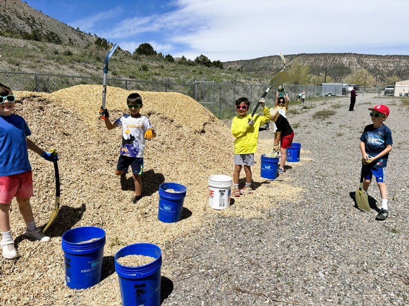 Students use buckets and shovels to move wood chips outside near a playground on a sunny day.