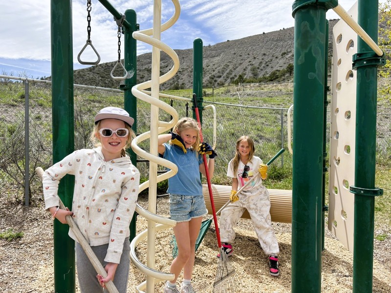 Three students smile while holding rakes in a playground covered with fresh wood chips