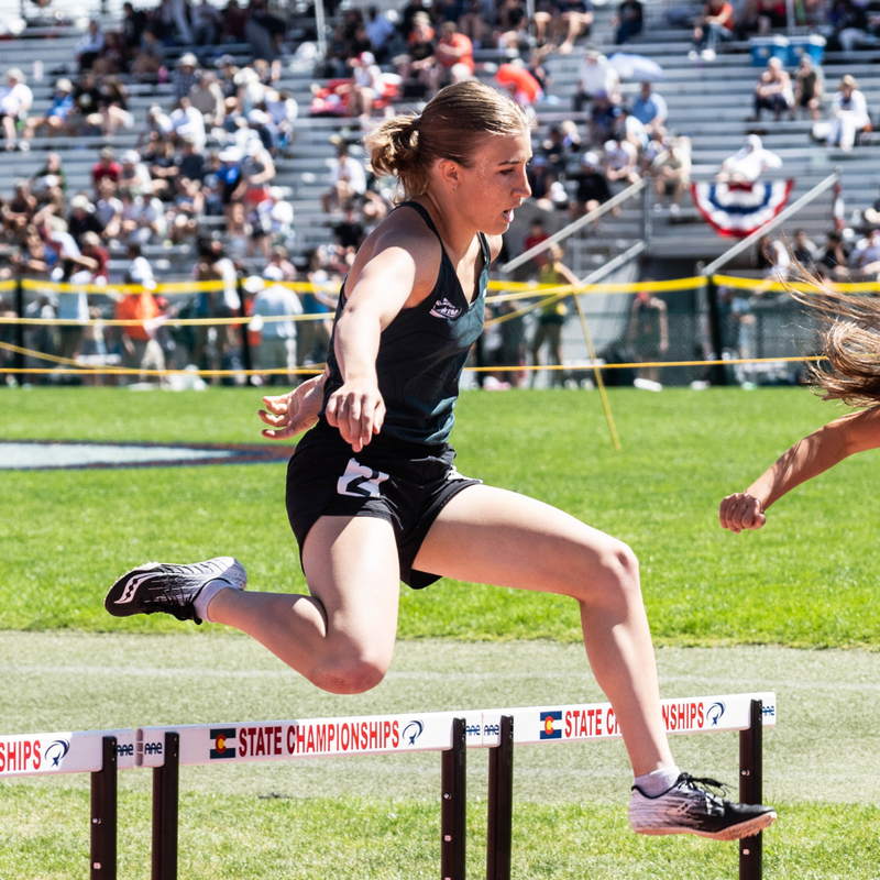Female track athlete clearing a hurdle during the Colorado State Track & Field Championships on a sunny day.