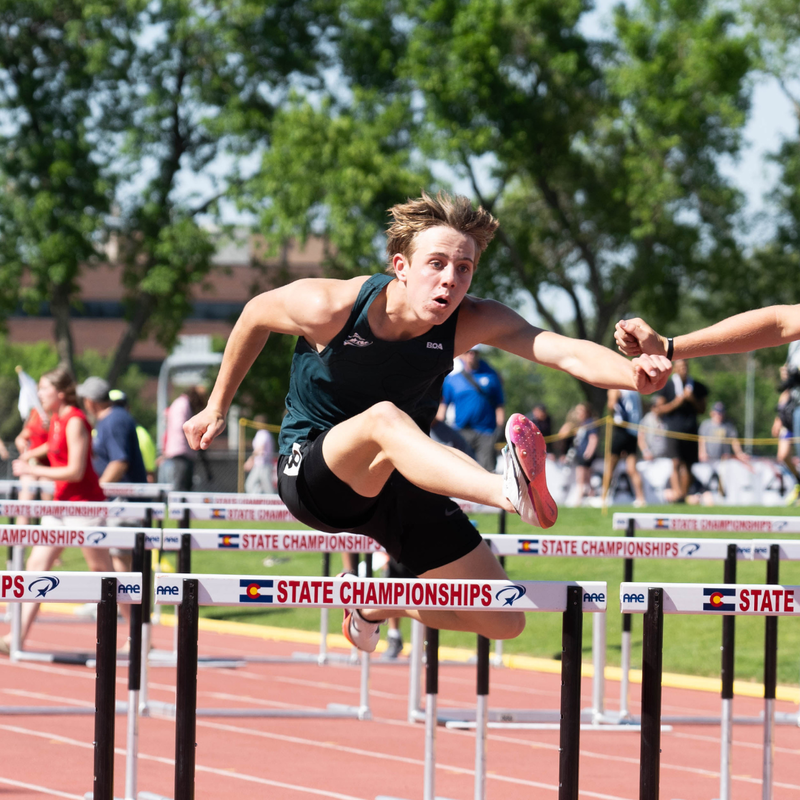Male track athlete clearing a hurdle during the Colorado State Track & Field Championships on a sunny day.
