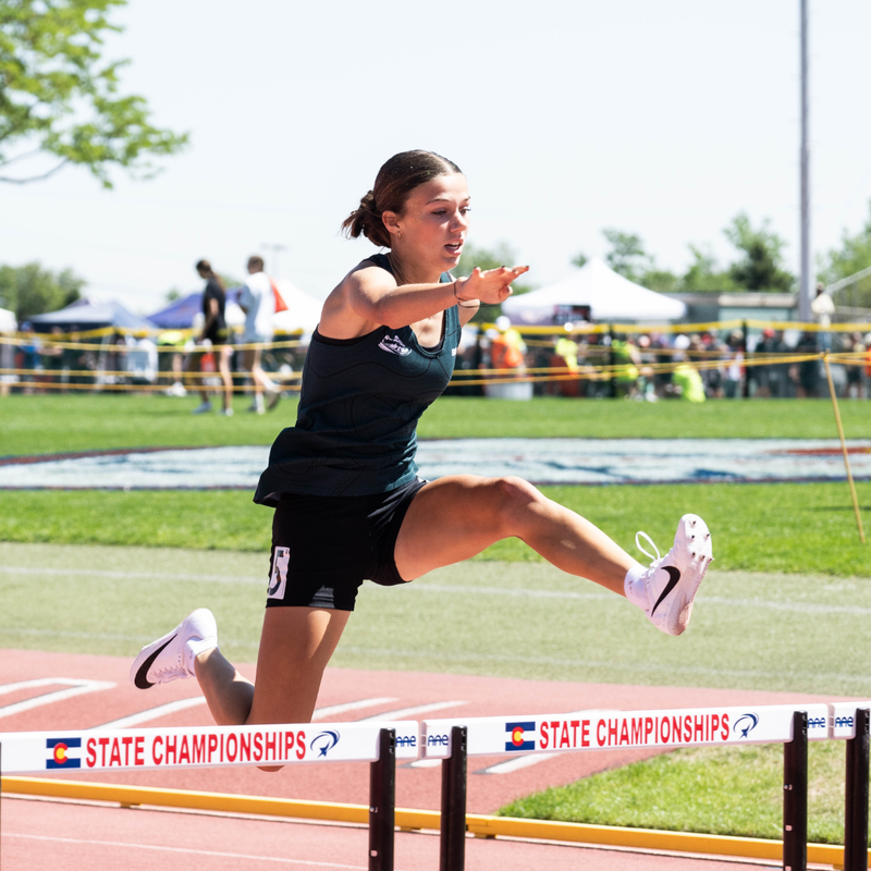 Female track athlete clearing a hurdle during the Colorado State Track & Field Championships on a sunny day.