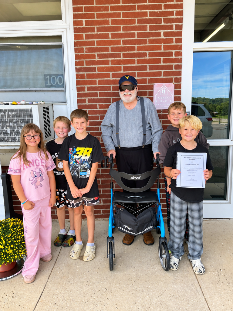 Five smiling elementary students pose outside a brick school building with an older man wearing a WV hat and standing behind a blue walker. One student holds a booklet titled Staunton-Parkersburg Turnpike in Ritchie County, WV. Yellow flowers are visible beside the group.