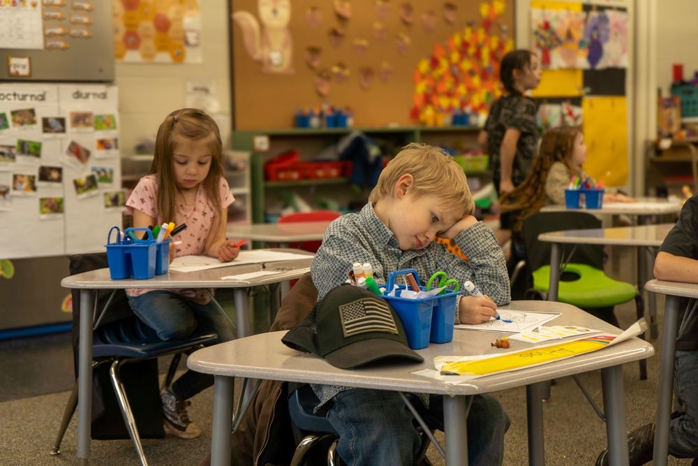 A little boy sits at a desk , coloring on a piece of paper. Other students work at other desks in the background.