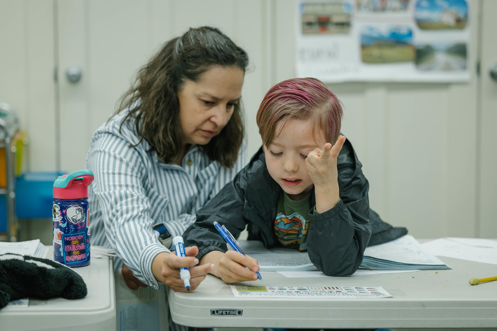 A teacher helps a young boy with his schoolwork at a desk.
