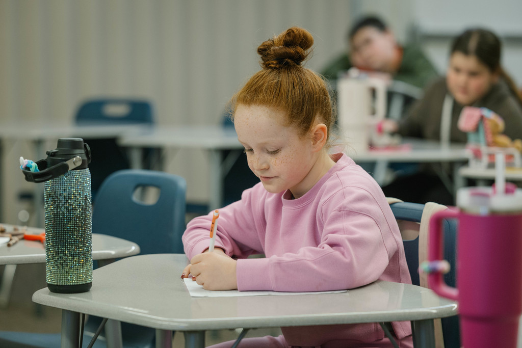 A young girl works at a desk, writing on a piece of paper.