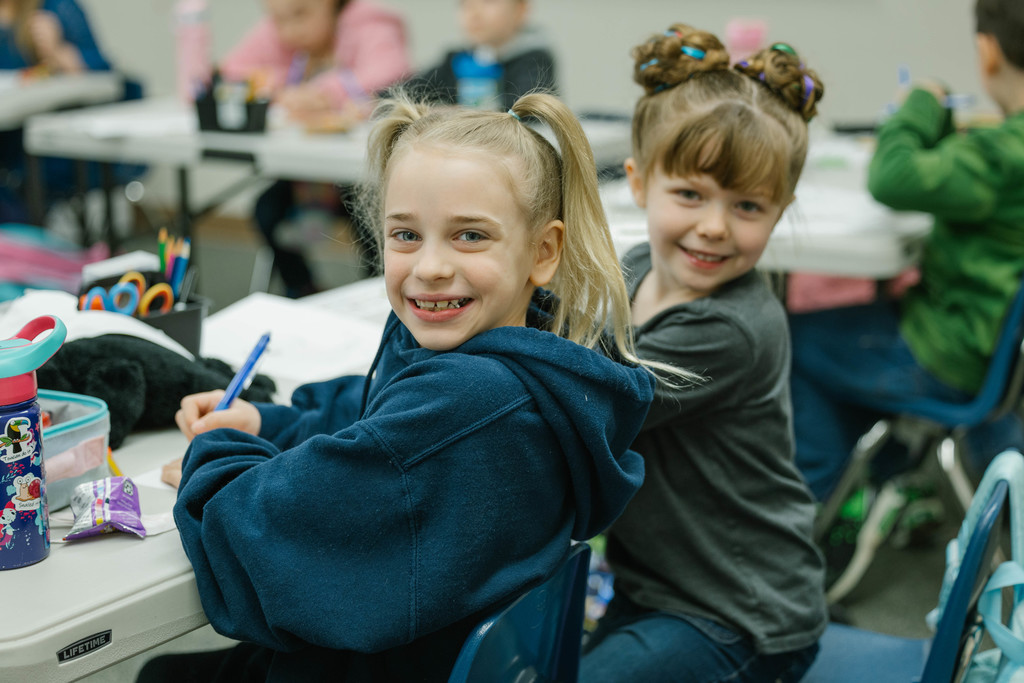 Two young girls smile at the camera as they work on schoolwork