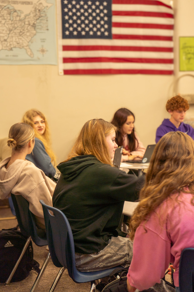 High school kids sit at desks with their laptops, chatting. An American flag is in the background.
