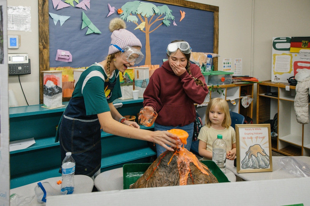 3 students smile and laugh, working together on a papermache volcano. 