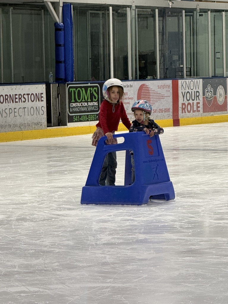 Two young girls, wearing helmets, use a push cart on ice to help them ice skate.