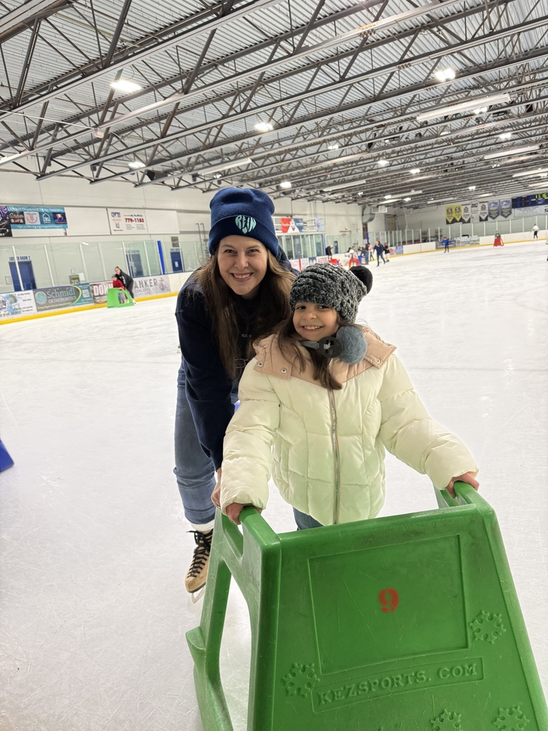 A woman and a little girl are ice skating. The girl is using a push cart to help her and the woman holds her from behind.