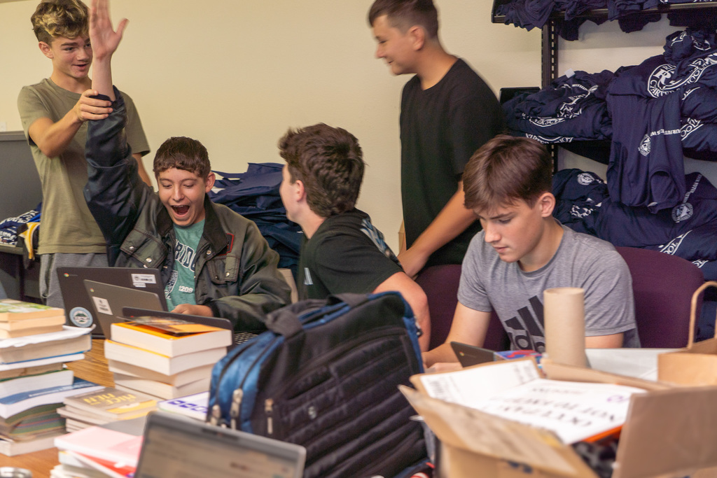 Five teen boys sit around computers and books. One holds his hand up in excitement and the others cheer him on.