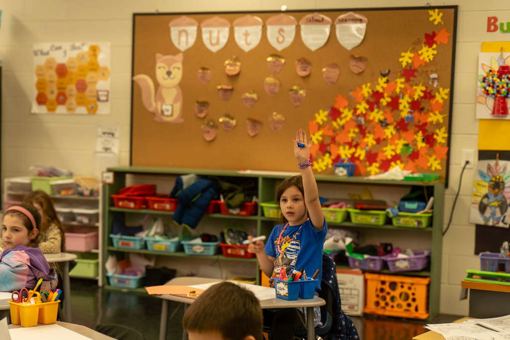 Young children sit at the desks in a classroom decorated for the fall. One young girl in a blue shirt raises her hand.