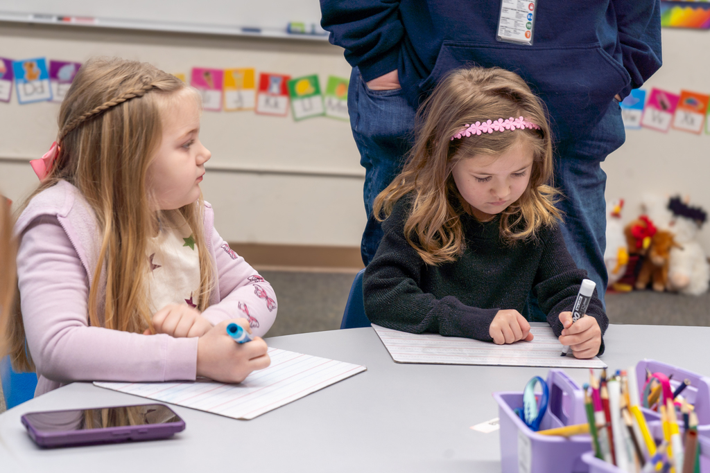 Two Kindergarten girls sit at a table, coloring with large markers on dry erase boards.