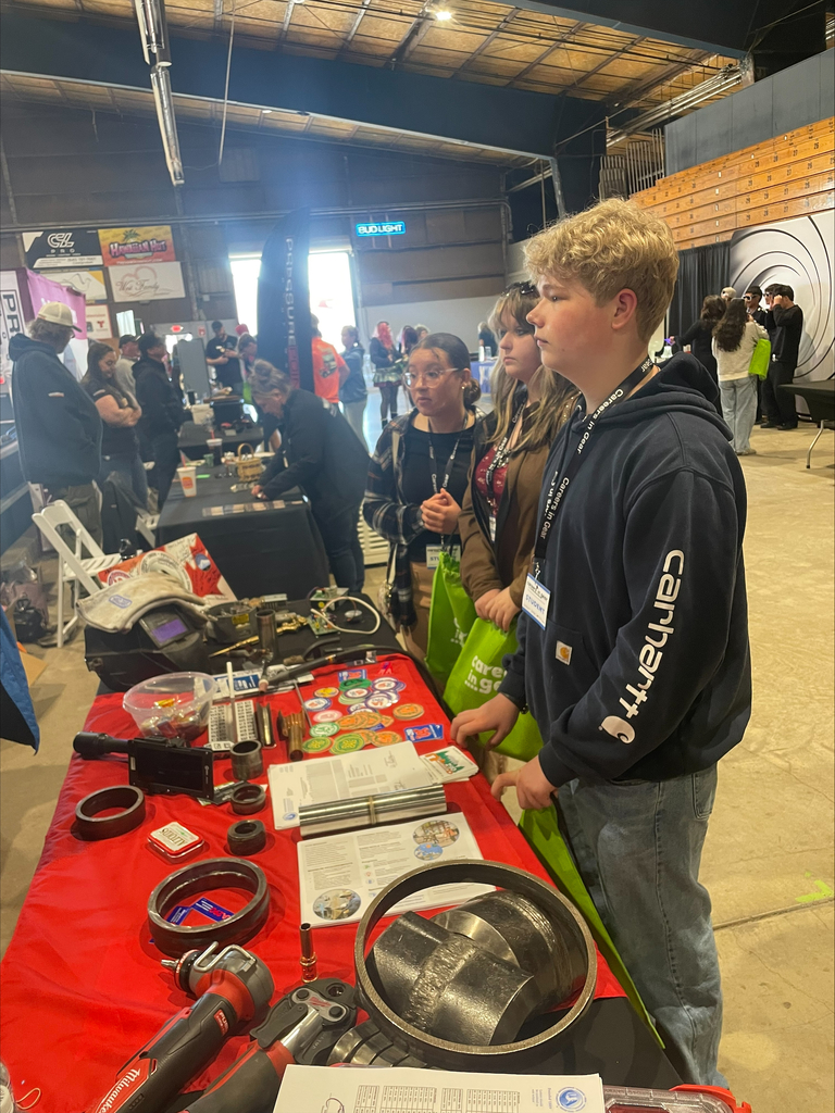 Two female and one male high school students stand in front of a table with several tools on it. There are more tables in the background with other people standing around them. 