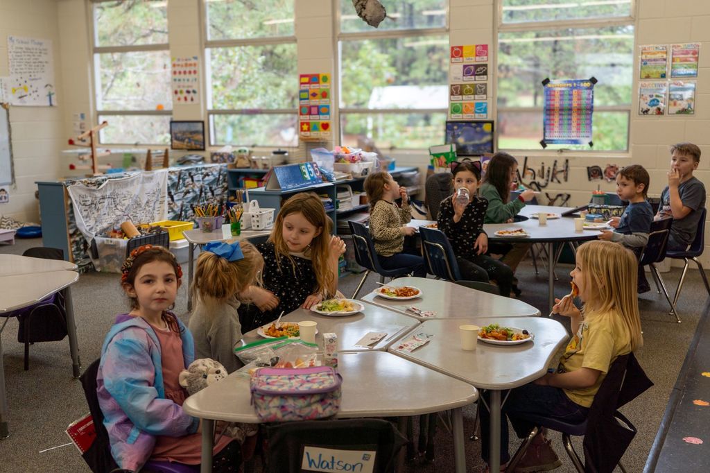 Elementary school students sit around tables with plates of lasagna in front of them.