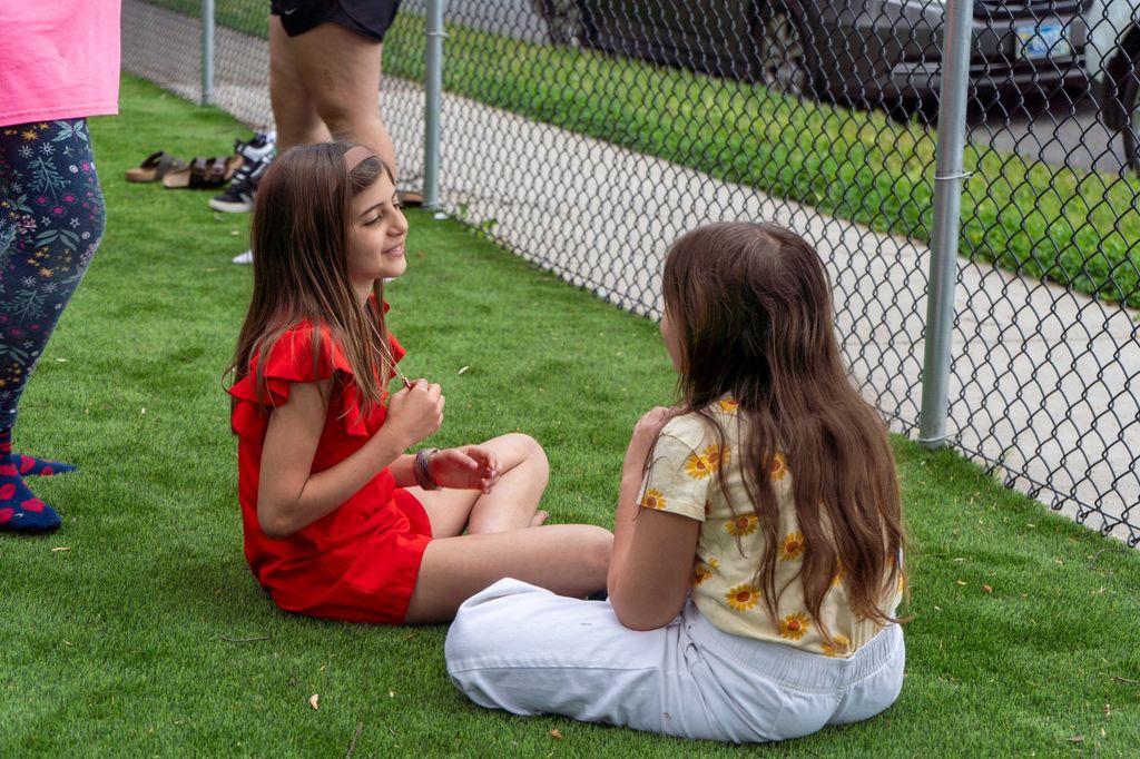 Two middle school girls sit on grass, talking to each other and smiling.