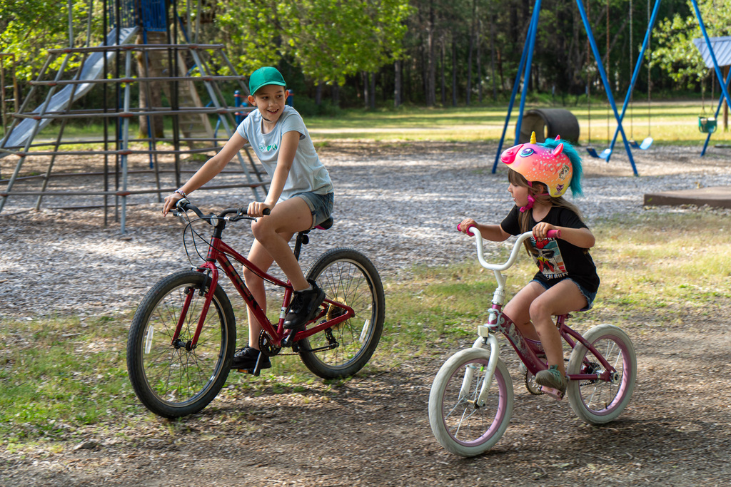 Two young girls ride bikes next to each other. They are smiling at each other.