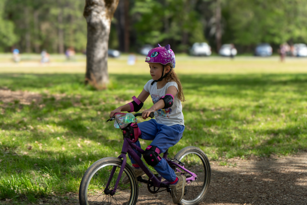 A young girl rides a bike. She is wearing knee and elbow pads. Her helmet is bright pink.