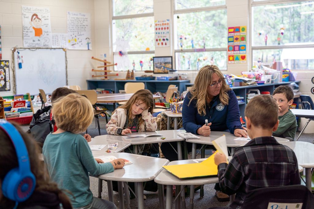 A female teacher sits at a circle of desks with elementary school students. They all work on papers in front of them. She points at one of the students' papers.