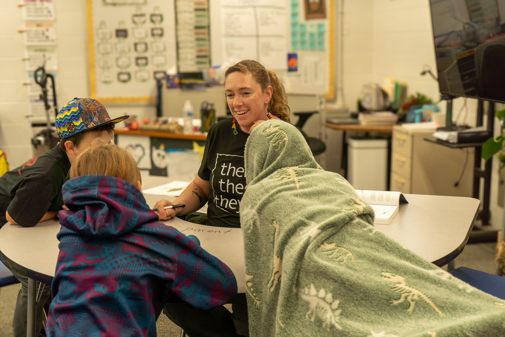 A female teacher sits in the middle of a U-shaped desk. Three students sit around her. She smiles at them. She is holding a pen.