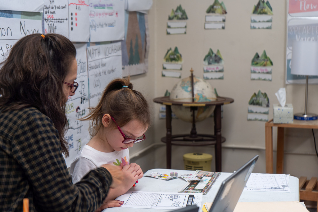 A female teacher sits at a table with a young girl in glasses. They work together on a sheet of paper.