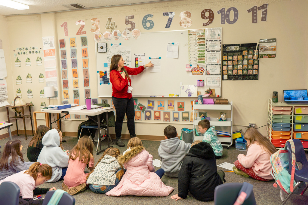 A female teacher points at a whiteboard. A class of elementary students sit on the ground around her, watching her.