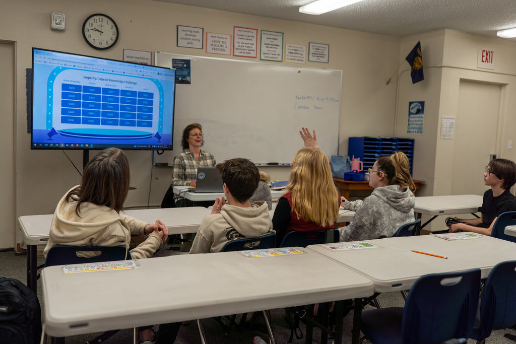 A female teacher sits in front of a class of middle school students. She has a game on a screen.