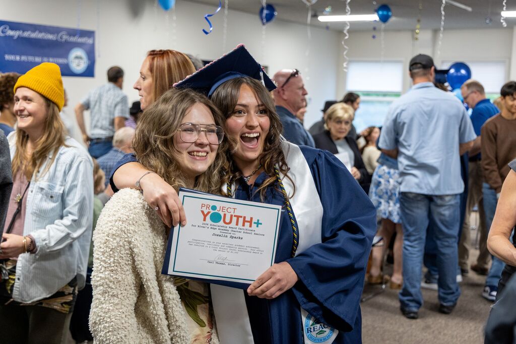A graduating senior girl in full regalia takes a picture with a teenage girl, holding a certificate in front of them. 