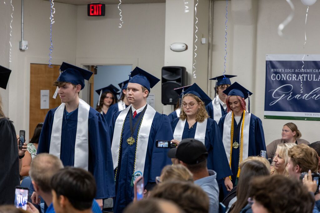 A line of high school seniors in graduation regalia walk down an aisle through a crowded room. 