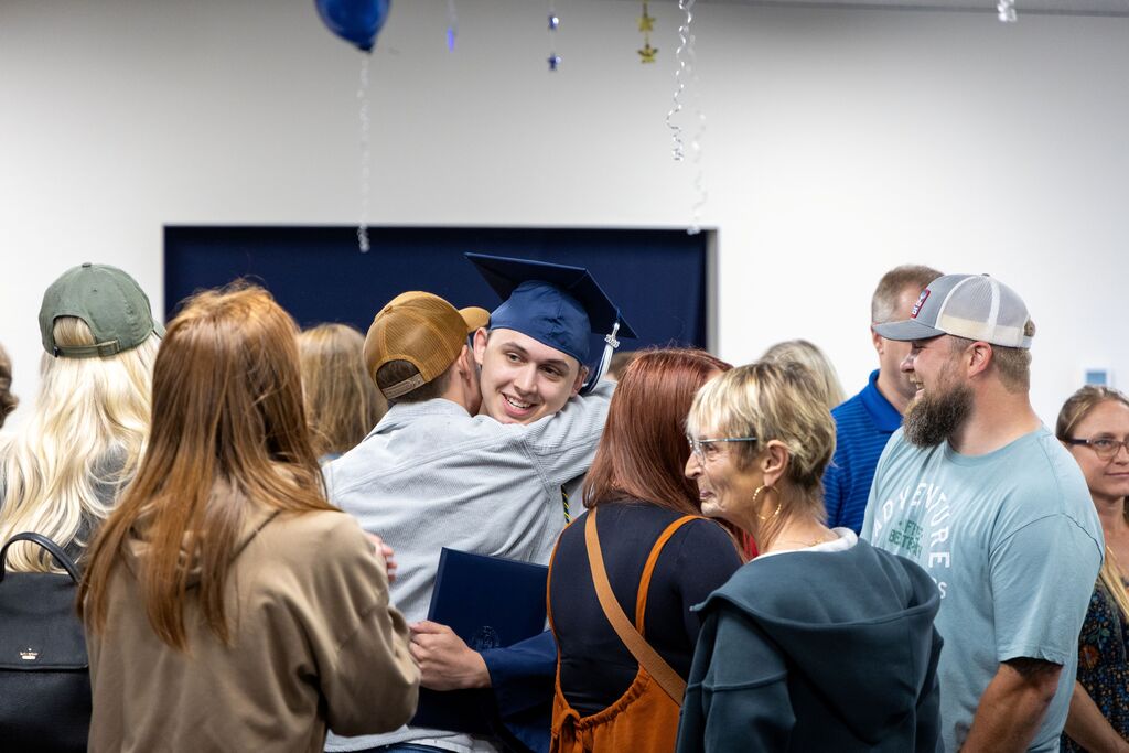 In a crowded room, a newly graduated young man hugs a man. 